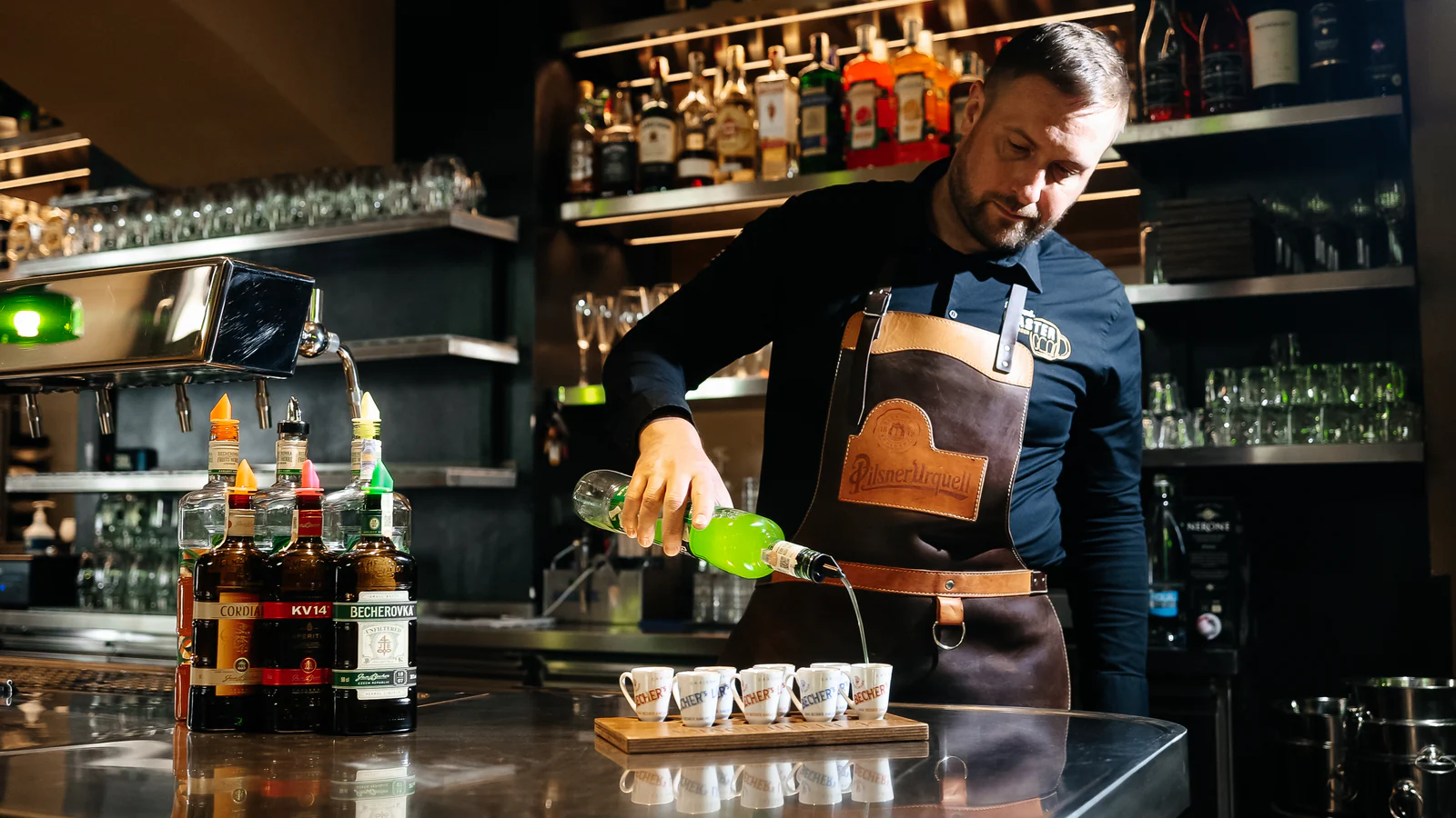 Barman nalévá zelený nápoj do malých skleniček v restauraci Plzeňka Carlsbad